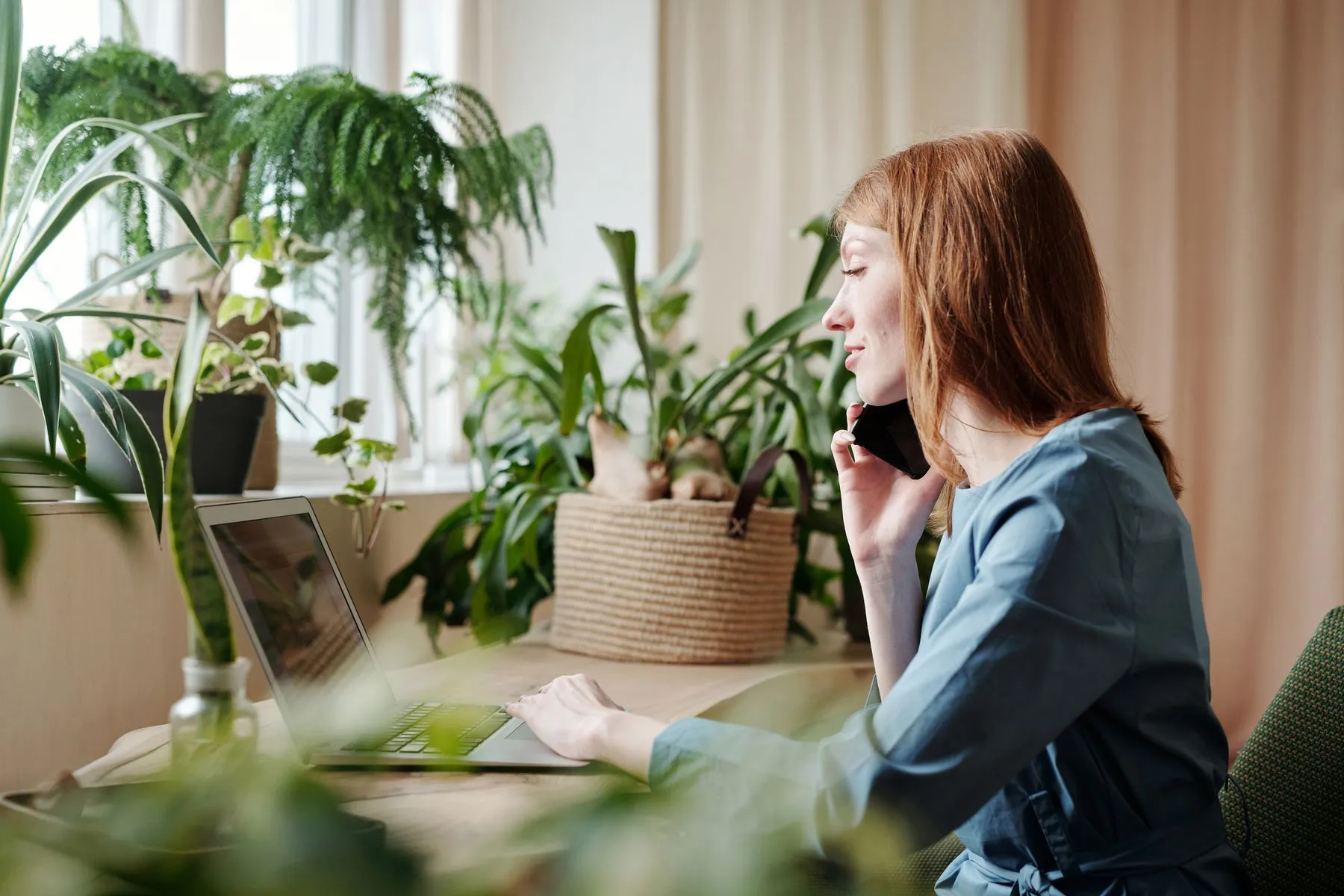 A woman on the phone at her laptop, surrounded by plants